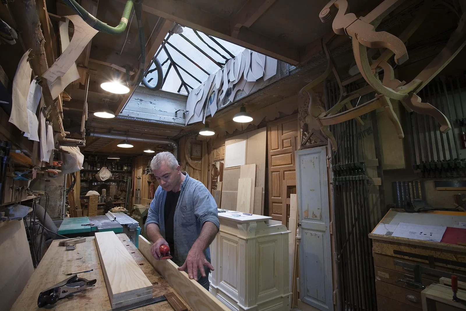 A carpenter measuring a long board at his bench in a busy workshop